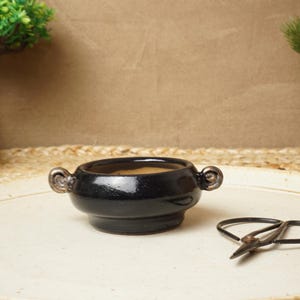 Può includere: Un vaso in ceramica nera con due manici, posto su una superficie bianca. Il vaso è vuoto e ha una leggera lucentezza. Un paio di piccole forbici da giardino nere sono anche sulla superficie.