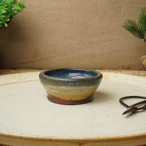 May include: A small, round, ceramic bowl with a blue, white and brown glaze. The bowl is sitting on a white ceramic tray.