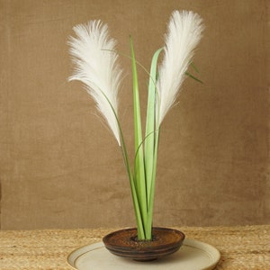 May include: A white pampas grass arrangement in a brown ceramic vase on a white plate. The vase has a textured surface and is sitting on a woven mat.