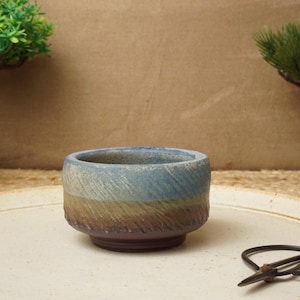 May include: A small, blue, brown and white ceramic pot with a textured surface. The pot is sitting on a white surface.