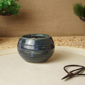 May include: A small, round, dark blue ceramic bowl with a glossy finish and horizontal grooved lines around the exterior. The bowl has a wide opening and a tapered base, showcasing a deep, mottled blue glaze with subtle variations in color.