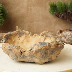 Peut inclure: Un pot en céramique rustique fait à la main avec une texture rugueuse et irrégulière. Le pot est de couleur brun clair avec des stries de glaçure brun foncé et bleue. Il est posé sur une assiette en céramique blanche.