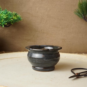 Op de afbeelding: Een kleine, donkerblauwe keramische bonsai pot met een getextureerd oppervlak. De pot staat op een crèmekleurige schaal, met een zwarte schaar in de buurt. Groene bonsai planten staan op de achtergrond.