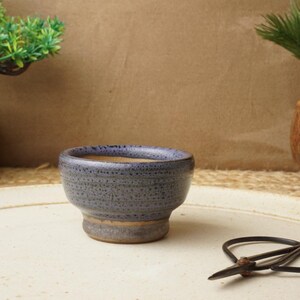 May include: A small, speckled blue ceramic bowl with a rounded shape and a slightly flared base. A pair of black scissors rests nearby on a cream-coloured surface. Two miniature bonsai trees in decorative pots are in the background.