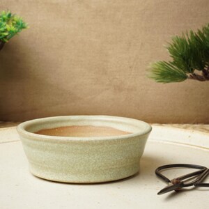 May include: A light green ceramic bowl with a slightly textured surface sits on a white tray. A pair of black bonsai shears are on the tray.