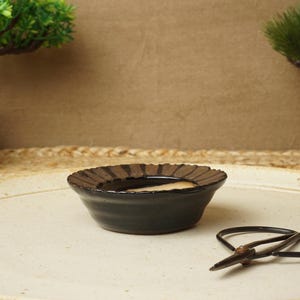Könnte beinhalten: Ein schwarzer Keramik-Bonsai-Topf mit brauner Glasur und einem strukturierten Rand. Der Topf steht auf einer weißen Oberfläche mit einer Bonsai-Schere.
