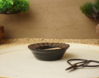 May include: A black ceramic bonsai pot with a brown glaze and a textured rim. The pot is sitting on a white surface with a pair of bonsai shears.