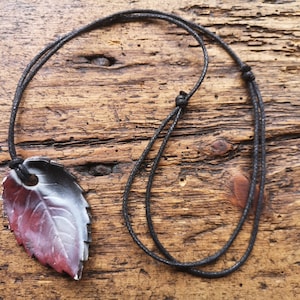Puede incluir: Un collar de cordón negro con un colgante de hoja roja, blanca y negra. El colgante de hoja tiene un patrón de vetas detallado.