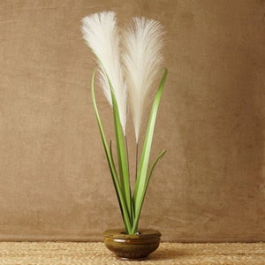 May include: A white pampas grass arrangement in a brown ceramic pot. The grass is tall and feathery, with green stems.