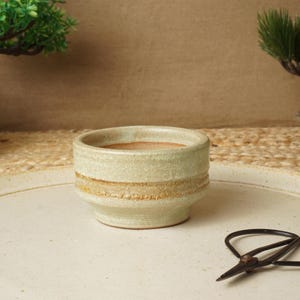 May include: A small, cylindrical ceramic pot with a textured, off-white glaze and a horizontal band of exposed, rough-textured tan clay around the center. The interior of the pot is smooth and light brown, featuring a thick rim and a tapered base.