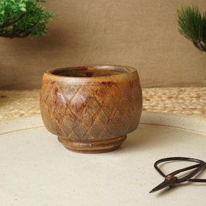 May include: A small, round, brown ceramic bowl featuring a textured diamond-patterned exterior. The bowl has a glazed finish with darker brown highlights and a distinct circular base. It is positioned on a flat, light-colored circular surface.
