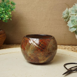 May include: A small, round ceramic pot with a textured, reddish-brown exterior and a dark brown interior. The pot sits on a cream-colored plate, with a pair of black scissors nearby. The pot is likely for plants or decorative use.