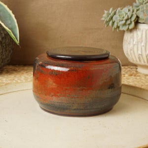 May include: A handcrafted ceramic jar with a lid. The jar features a gradient of colors, transitioning from red at the top to dark blue at the base. It sits on a cream-colored plate, suggesting a decorative or storage item.