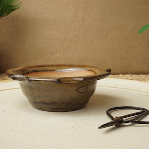 May include: A small, round, brown ceramic bonsai pot with a wide rim and a slightly textured surface. The pot is on a large, off-white ceramic plate. A pair of small, black scissors rests nearby.