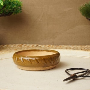 May include: A small, round, green ceramic pot with a textured surface. The pot is sitting on a white surface next to a pair of bonsai shears.