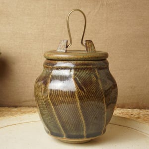 Op de afbeelding: Een bruine keramische pot met een deksel en een messing handvat. De pot heeft een gestreept patroon en een glanzende afwerking.