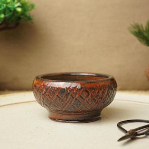 May include: A small, round, terracotta-coloured ceramic pot with a geometric pattern. The pot has a dark brown glaze and a shiny finish.