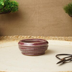 Peut inclure: Un petit plat rond en céramique avec un glaçage violet foncé et un bord blanc. Le plat est posé sur une surface blanche.