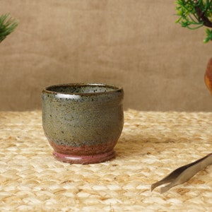 May include: A small, grey and brown ceramic cup with a pink rim. The cup is sitting on a woven mat.