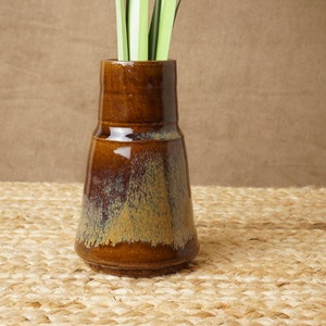 May include: A brown ceramic vase with a speckled glaze. The vase has a wide base and a narrow neck. It is sitting on a woven mat.