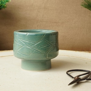 May include: A teal ceramic planter with a geometric pattern. The small, round planter sits on a pedestal base. A pair of black scissors rests on the surface in front of the planter. The background is a neutral tan color.