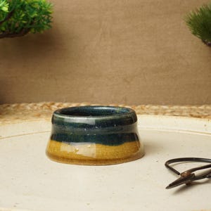 May include: A small, round ceramic dish with a blue and yellow glaze. The dish is sitting on a white surface.