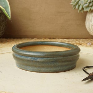 Könnte beinhalten: Kleiner, runder Bonsai-Topf aus Keramik mit blaugrüner Glasur. Der Topf hat ein niedriges Profil mit einer weiten Öffnung und einer strukturierten Außenseite. Der Innenraum ist hellbraun. Eine schwarze Schere liegt auf der Oberfläche.