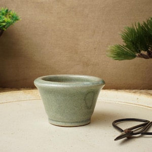 Może przedstawiać: Mały, jasnozielony ceramiczny garnek z zaokrąglonym dnem i lekko rozszerzającym się brzegiem. Garnek stoi na białym ceramicznym talerzu z małymi nożyczkami ogrodowymi.