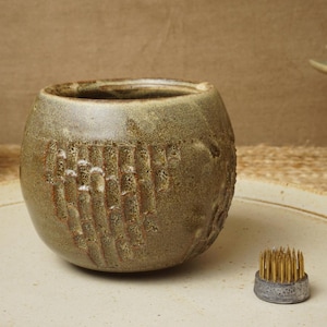 May include: A green ceramic vase with a textured surface. The vase has a round shape and a wide opening. A small metal flower frog is in the foreground.