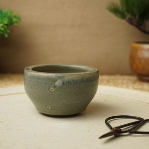 以下が含まれることがあります： 斑点のある釉薬が施された、小さな薄い緑色の陶器のボウル。ボウルは白い皿の上に置かれ、小さな黒いハサミが置かれている。