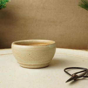 以下が含まれることがあります： 小さなベージュ色の陶器のボウルが、大きなオフホワイトの陶器の皿の上に置かれています。黒いハサミが皿の上に置かれています。背景はニュートラルなタン色で、両側に小さな鉢植えの植物があります。