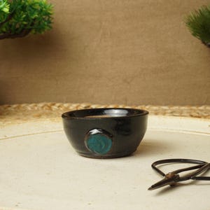 Könnte beinhalten: Eine kleine schwarze Keramikschale mit einem türkisfarbenen Kreisdesign. Die Schale steht auf einem weißen Tablett mit einer braunen geflochtenen Matte darunter. Auf dem Tablett liegt auch eine schwarze Bonsai-Schere.