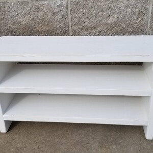 K&ouml;nnte beinhalten: Eine wei&szlig;e Holzbank mit zwei Regalen. Die Bank hat eine rechteckige Oberseite und ein schlichtes Design. Die Regale sind gleichm&auml;&szlig;ig verteilt. Die Bank ist wei&szlig; lackiert. Abmessungen: ca. 90 cm lang, 30 cm hoch und 30 cm tief.
