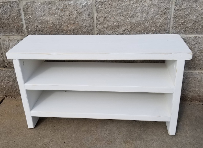 K&ouml;nnte beinhalten: Eine wei&szlig;e Holzbank mit zwei Regalen. Die Bank hat eine rechteckige Oberseite und ein schlichtes Design. Die Regale sind gleichm&auml;&szlig;ig verteilt. Die Bank ist wei&szlig; lackiert. Abmessungen: ca. 90 cm lang, 30 cm hoch und 30 cm tief.