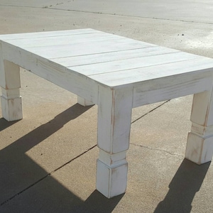 May include: A rectangular white wooden coffee table with a distressed finish. The table has a slatted top and four thick, square legs. The table is approximately 48 inches long, 24 inches wide, and 18 inches high.