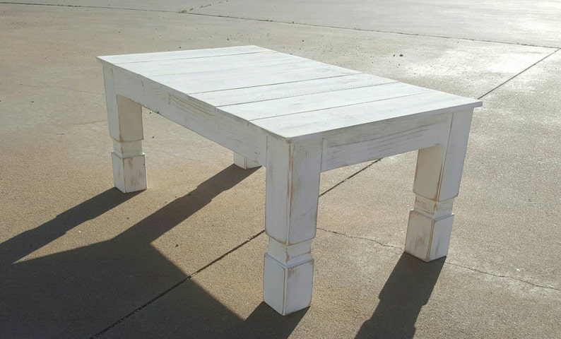 May include: A rectangular white wooden coffee table with a distressed finish. The table has a slatted top and four thick, square legs. The table is approximately 48 inches long, 24 inches wide, and 18 inches high.