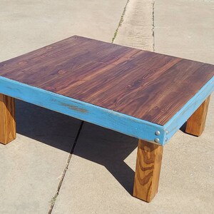 Peut inclure: Une table basse en bois faite &agrave; la main avec un dessus teint&eacute; fonc&eacute; et une bordure bleu clair. La table a quatre pieds en bois naturel robustes et est construite avec des attaches m&eacute;talliques visibles. La table mesure environ 76 cm de long, 61 cm de large et 41 cm de haut.