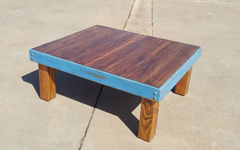 Peut inclure: Une table basse en bois faite &agrave; la main avec un dessus teint&eacute; fonc&eacute; et une bordure bleu clair. La table a quatre pieds en bois naturel robustes et est construite avec des attaches m&eacute;talliques visibles. La table mesure environ 76 cm de long, 61 cm de large et 41 cm de haut.