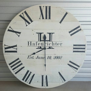 May include: A large, round, white wooden clock with black Roman numerals and hands. The clock face features the name "Hafenrichter" and the date "Est. June 10, 2002". The clock is a decorative piece for home decor.