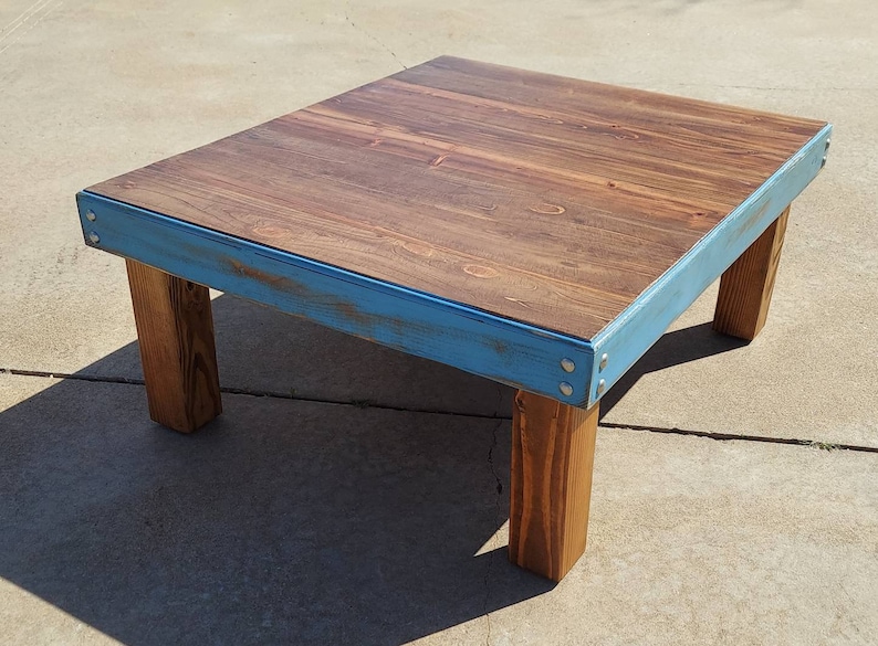 Peut inclure: Une table basse en bois faite &agrave; la main, avec un plateau rectangulaire et quatre pieds robustes. Le plateau est teint&eacute; d'un brun riche, accentu&eacute; par une bordure bleu clair. La table mesure environ 76 cm de long, 51 cm de large et 41 cm de haut.