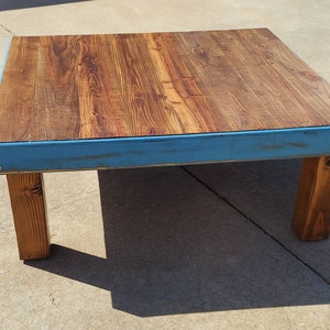Peut inclure: Une table basse carr&eacute;e en bois avec un dessus en grain de bois naturel et une bordure peinte en bleu vieilli. La table est soutenue par quatre pieds carr&eacute;s en bois robustes. La table mesure environ 91 cm sur 91 cm.
