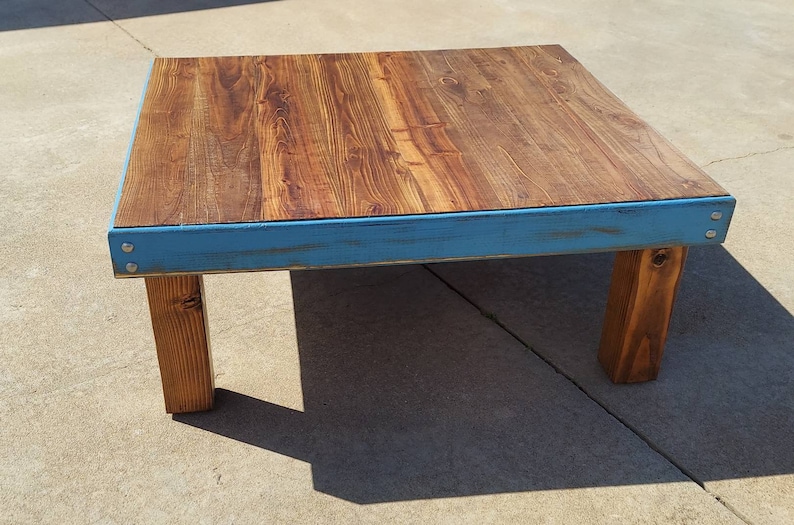 Peut inclure: Une table basse carr&eacute;e en bois avec un dessus en grain de bois naturel et une bordure peinte en bleu vieilli. La table est soutenue par quatre pieds carr&eacute;s en bois robustes. La table mesure environ 91 cm sur 91 cm.