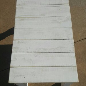 May include: A rectangular, white-painted wooden coffee table. The tabletop is constructed from horizontal planks, showing the wood grain. The table has a distressed, weathered appearance, with visible knots and imperfections. The table is supported by four short, square legs.