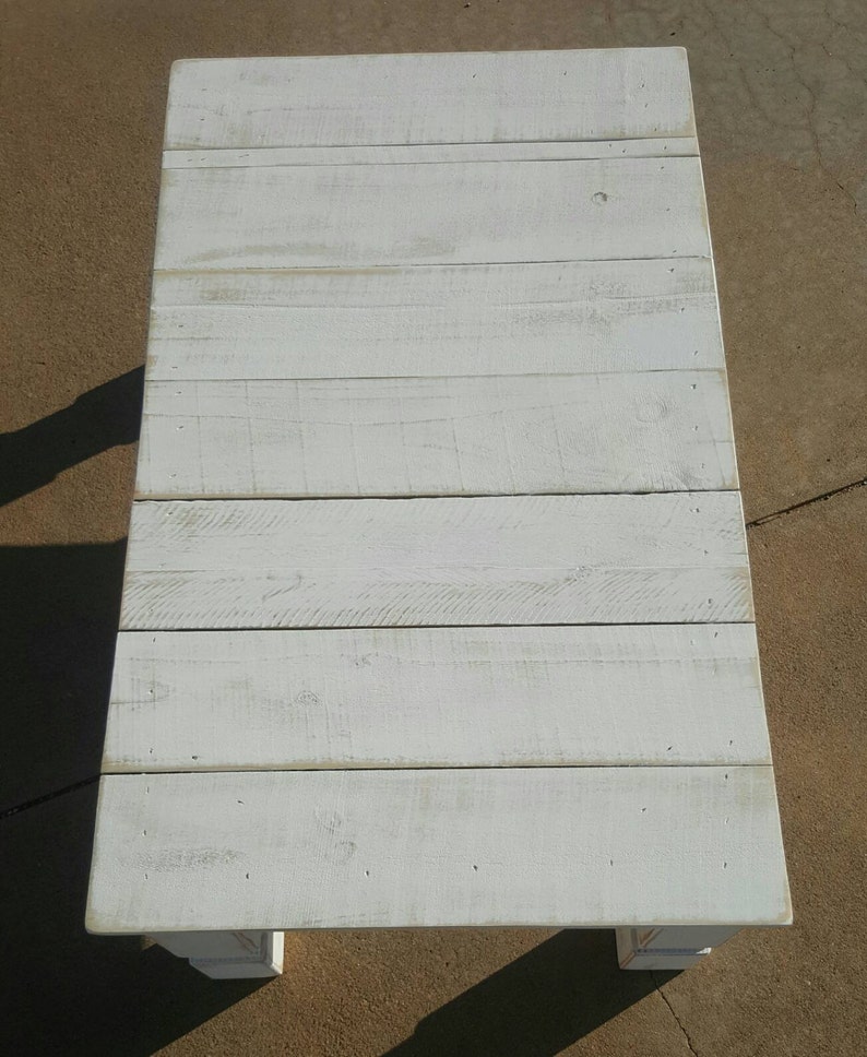 May include: A rectangular, white-painted wooden coffee table. The tabletop is constructed from horizontal planks, showing the wood grain. The table has a distressed, weathered appearance, with visible knots and imperfections. The table is supported by four short, square legs.