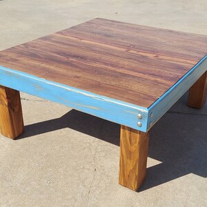 Peut inclure: Table basse en bois faite &agrave; la main, avec un plateau rectangulaire et quatre pieds. Le plateau pr&eacute;sente un veinage de bois fonc&eacute;, encadr&eacute; par un bord peint en bleu clair. Les pieds sont en bois naturel, pour un look rustique et moderne. Mesure environ 91 cm de long.