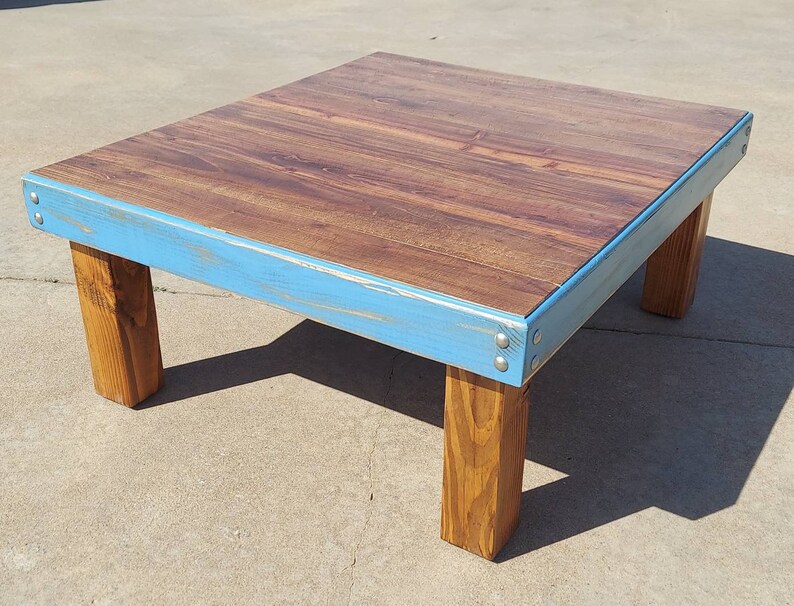 Peut inclure: Table basse en bois faite &agrave; la main, avec un plateau rectangulaire et quatre pieds. Le plateau pr&eacute;sente un veinage de bois fonc&eacute;, encadr&eacute; par un bord peint en bleu clair. Les pieds sont en bois naturel, pour un look rustique et moderne. Mesure environ 91 cm de long.