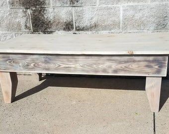 May include: A rectangular wooden coffee table with a light gray finish. The table has a plank-style top and four angled legs. The wood grain is visible on the table's apron. The table is approximately 48 inches long, 24 inches wide, and 16 inches high.