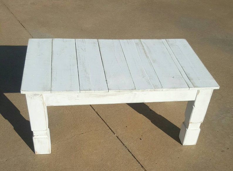 May include: A rectangular white wooden coffee table with a distressed finish. The table has a slatted top and sturdy, square legs. The table is approximately 48 inches long, 24 inches wide, and 18 inches high.