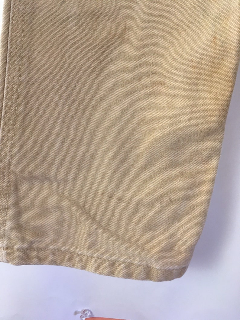 May include: Close-up of tan-colored canvas fabric, showing the texture and stitching details. The fabric has some minor discoloration and wear, suggesting it is a well-used item. The bottom hem is visible.