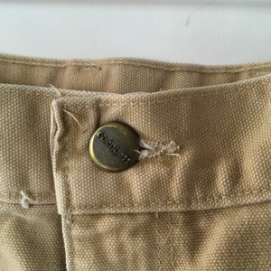 May include: Close-up of tan-colored fabric with a metal button. The button is circular with the brand name "Carhartt" embossed on it. The fabric appears to be a durable canvas material, commonly used in workwear.